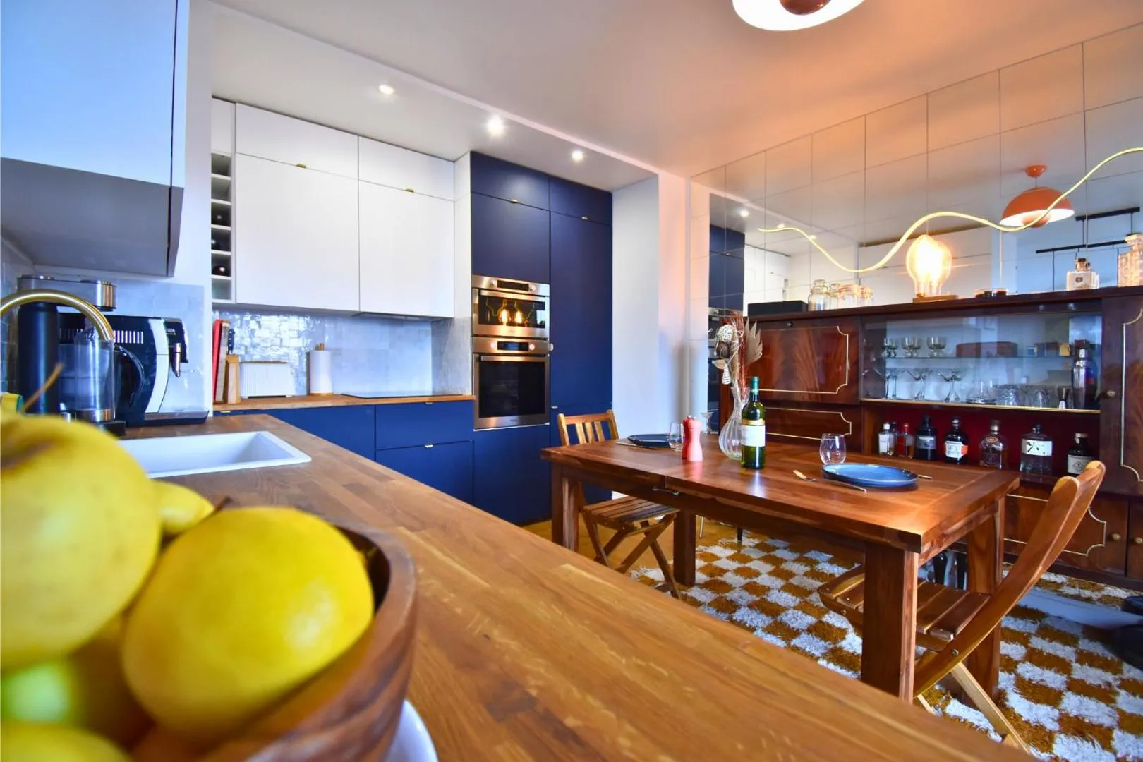 Cuisine après : façades bleu marine, plan de travail bois, îlot avec citrons, table en chêne, buffet ancien, suspension orange, tapis à carreaux moutarde