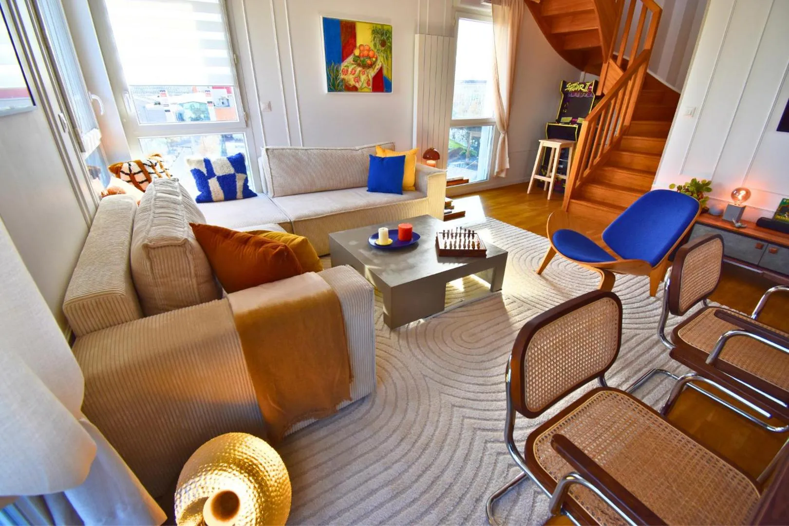 Salon duplex après : canapé en lin beige, fauteuils en rotin, fauteuil bleu accent, escalier bois, luminaire globe