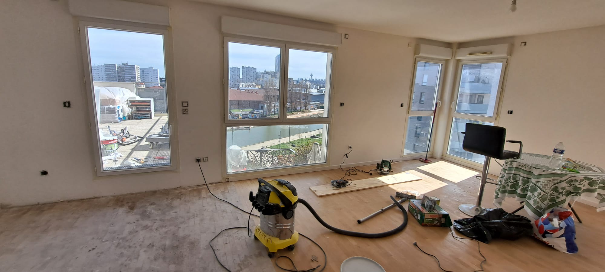Salon brut pendant les travaux : parquet, outils, aspirateur, grande baie vitrée vue canal