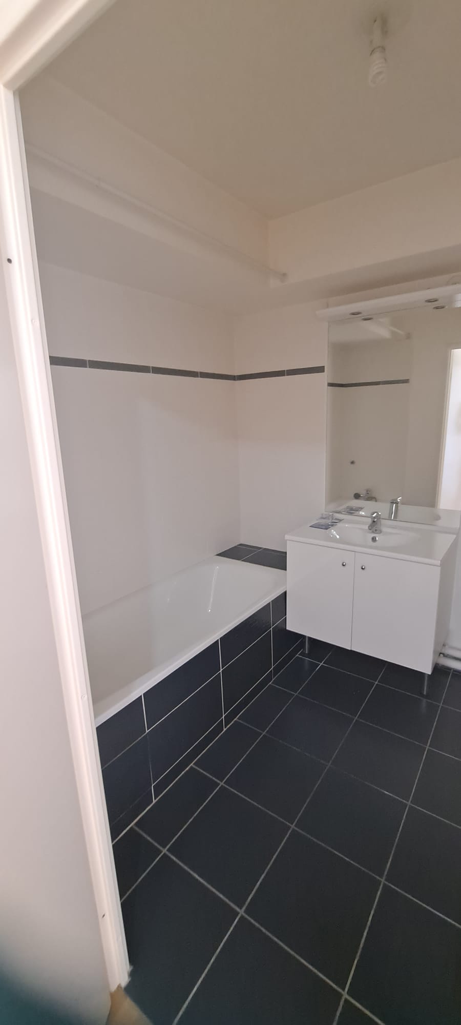 Salle de bain avant : carrelage noir, baignoire standard, meuble blanc générique