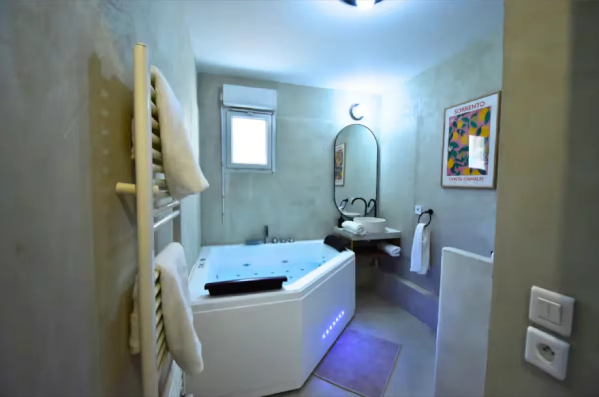 Salle de bain après : béton ciré gris, baignoire balnéo à jets avec LED bleues, miroir oval, serviettes roulées, affiche colorée