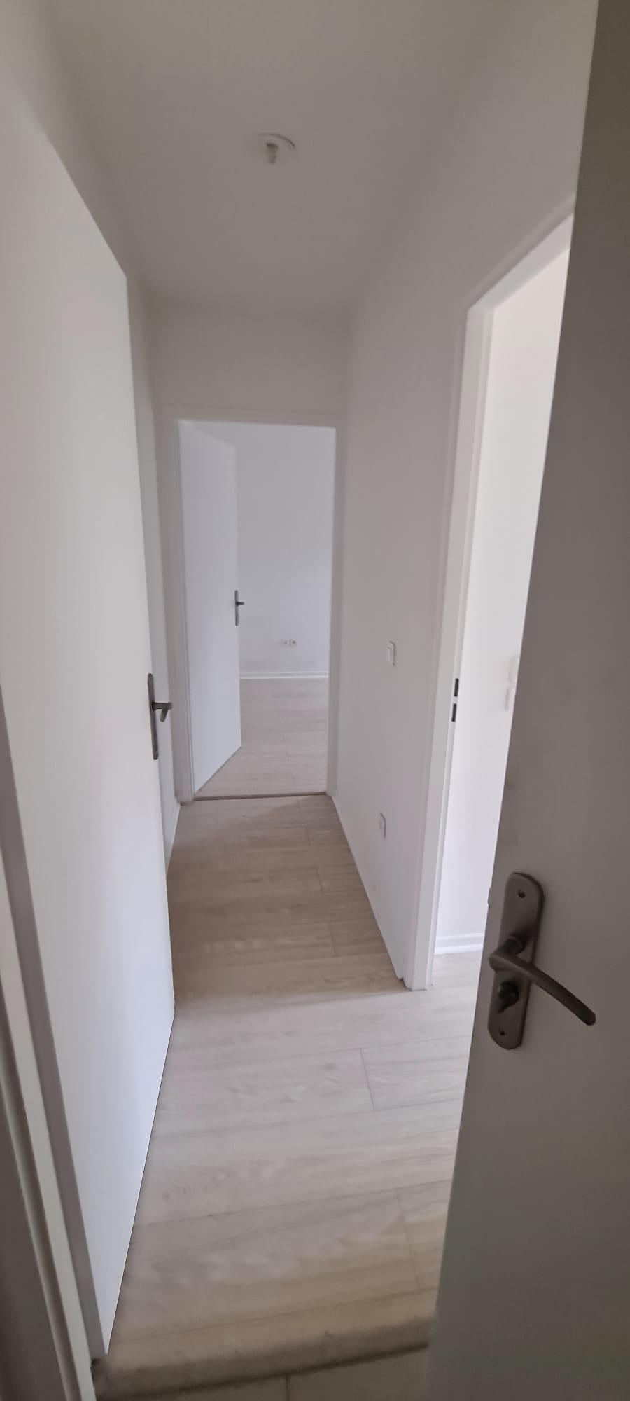 Couloir vide : murs blancs, parquet clair, porte, rien d'autre