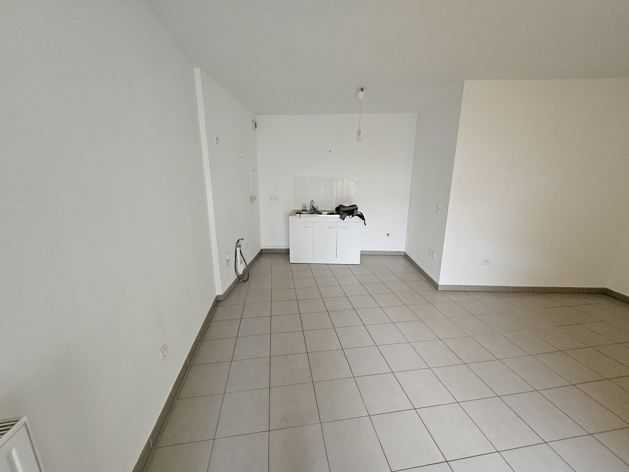 Séjour vide : carrelage gris, murs blancs, mini-kitchenette dans le fond, ampoule nue au plafond