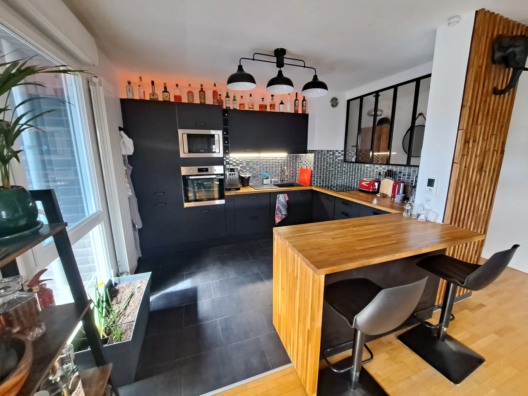 Cuisine après rénovation : façades noires, plan de travail bois, verrière, bar et éclairage chaleureux